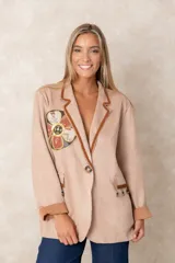 Blazer de corte recto en color beige, con solapas decoradas con ribete tejido en tono marrón. Presenta un aplique circular bordado con motivos florales y detalles de cuentas en el pecho, además de bolsillos frontales con flecos de cuentas colgantes. Posee cierre de un solo botón y mangas con puños vueltos en un tono más oscuro.