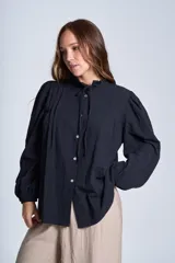 Camisa de color azul con cuello alto decorado con volados y lazo de ajuste. Presenta tablas decorativas en los hombros, cierre frontal con botones de nácar y mangas largas con puños elásticos que generan un efecto abullonado.