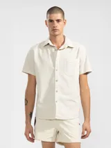 Camisa de manga corta color crema con textura tipo waffle, cuello clásico, cierre con botones y bolsillo de parche en el pecho. Corte recto y holgado.