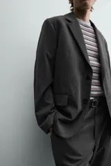 Blazer oversized fit de hombre, color gris oscuro con rayas verticales finas. Cuello con solapas de muesca, manga larga con puño abotonado, bolsillos de solapa en la cadera y bolsillo interior. Cierre frontal de un botón y abertura en el centro de la espalda.