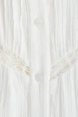 Vestido túnica corto de algodón con efecto arrugado y diseño holgado. Cuenta con inserciones de encaje, cuello redondo y cierre frontal de botones. Presenta mangas globo largas con puños elásticos, cinturón fino extraíble para ajustar la cintura y costura fruncida en el bajo con volados.