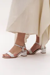 Sandalia de cuero color plateado, con tira ancha en la parte delantera y tira fina con hebilla alrededor del tobillo. Tiene un tacón cuadrado de altura media.