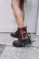 Botas cortas de cuero color marrón oscuro con suela de goma negra gruesa y dentada. Cuentan con un cierre lateral de cremallera de color rojo brillante y un tirador de tela rojo en la parte trasera superior.