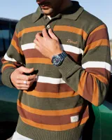 Buzo tejido de punto con diseño de rayas horizontales en tonos verde, marrón y blanco. Presenta cuello tipo polo con cierre de tres botones, mangas largas con puños acanalados y logo de la marca bordado en el pecho. Incluye una etiqueta decorativa en la parte inferior.