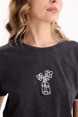 Remera crop negra con efecto acid wash, mangas cortas con costuras decorativas en blanco y bordado frontal de un florero con flores.