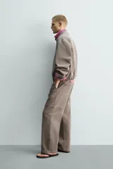 Pantalón de corte relajado en color marrón con efecto lavado. Presenta bolsillos frontales tipo cargo, cierre con cremallera y botón, y una línea horizontal de tachas metálicas a tono en la parte superior de las piernas.