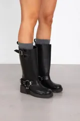 Bota de caña alta estilo biker, confeccionada en cuero sintético negro. Presenta una suela track con taco cuadrado, una hebilla ajustable en la parte superior de la caña y un arnés decorativo en el tobillo con argolla metálica y remaches.