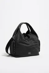 Bolso tipo hobo o tote de nylon negro, con asas de mano cortas y una correa bandolera ajustable y desmontable de tejido negro. Presenta bolsillos laterales con cierre de cremallera y el logo de Bimba y Lola en blanco en la parte frontal inferior.