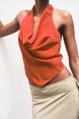 Top de color naranja vibrante con cuello halter que se anuda en la espalda, creando un escote drapeado o fluido en el frente. El bajo del top tiene un corte en pico con tiras finas colgantes.