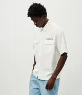 Camisa de manga corta con corte oversized, confeccionada en tejido texturizado de viscosa y poliéster. Presenta cuello clásico, cierre frontal con botones, dos bolsillos frontales con solapa y detalle de lettering bordado en el pecho.