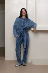 Mameluco enterito de jean con efecto nevado vintage, de corte holgado, mangas largas, cuello mao, cierre frontal con botones y lazo ajustable en la cintura.