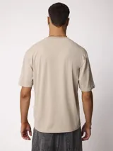 Remera de manga corta color gris piedra, de corte oversize y cuello redondo, con pequeño parche bordado en el dobladillo inferior.
