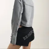Short de denim negro con diseño de tiro bajo, bolsillos traseros con apliques de strass formando la inscripción "BROOKLYN" y terminaciones deshilachadas.