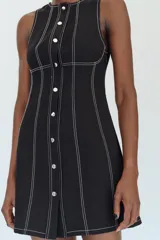 Vestido mini negro con cuello redondo y diseño sin mangas, con pespuntes en contraste y botones decorativos frontales.