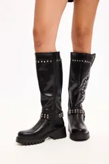 Bota de caña media en cuero sintético negro, con suela track, cierre lateral y detalle de tachas metálicas en la parte superior y en una correa decorativa en el tobillo.