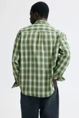 Camisa de manga larga de corte relajado, confeccionada en mezcla de algodón suave con estampado a cuadros en tonos verdes y blancos. Presenta cuello inglés, cierre frontal de botones, un bolsillo aplicado en el pecho y dobladillo redondeado.