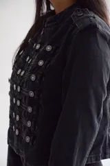 Chaqueta de estilo militar confeccionada en denim negro con efecto lavado. Presenta un diseño estructurado con cierre frontal de cremallera, cuello mao y múltiples botones metálicos decorativos dispuestos en filas horizontales. Incluye detalles de charreteras en los hombros y puños con botones.
