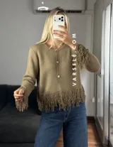 Cárdigan de tejido de punto color crema, con cuello redondo, mangas largas y cierre frontal con botones metálicos. Presenta detalles de flecos en los puños y en el ruedo.