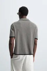 Camisa de punto de manga corta con cuello bowling y cierre frontal de botonadura. Presenta un patrón de rayas horizontales finas en blanco y negro, con ribetes negros en cuello, puños y bajo.