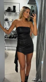 Vestido corto strapless de tela satinada negra, con diseño ajustado y fruncido lateral. Presenta un detalle de caída asimétrica en uno de los costados.