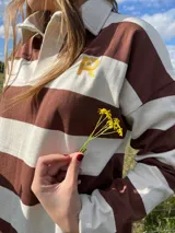 Sweater estilo rugby de manga larga con diseño de rayas horizontales anchas en color chocolate y crudo. Presenta cuello tipo polo con cartera de botones y un logo bordado en color amarillo en el pecho.