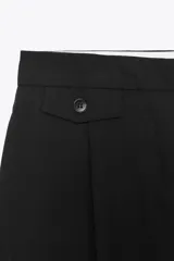 Pantalón de vestir marrón oscuro, tiro alto, con pernera ancha y pinzas frontales. Presenta un bolsillo con tapeta en la parte superior delantera.