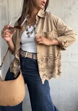 Sobrecamisa de gamuza color beige, con cuello camisero, cierre frontal con botones metálicos y bolsillo en el pecho. Presenta un diseño con calados florales y bordes ondulados en la parte inferior.
