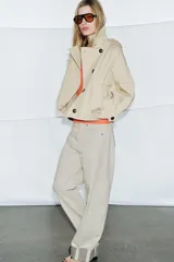 Trench corto color verde militar, con cuello solapa y capucha. Tiene manga larga con trabillas y botón en los puños, bolsillos delanteros y bajo asimétrico. Cierre frontal con botones. El tejido es repelente al agua.