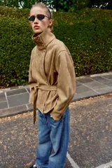 Chaqueta de gamuza sintética color marrón, con cuello subido, cierre frontal cruzado con botones y mangas largas anchas. Cuenta con bolsillos delanteros y cinturón ajustable del mismo tejido.