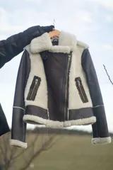 Campera tipo gamulán de cuero genuino color marrón oscuro, con interior y detalles en piel sintética color crema. Presenta cierre frontal cruzado, bolsillos laterales con cierre y puños con borde de piel.