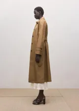 Chaqueta larga de algodón color marrón tostado, estilo gabardina, con cuello camisero, cierre frontal oculto con botones, mangas largas con presillas en los puños y cinturón ajustable con hebilla oscura.