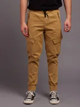 Pantalon tipo cargo color tostado, confeccionado en sarga. Presenta cintura elástica con cordón de ajuste, bolsillos laterales con solapa y puños elásticos en los tobillos.