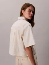 Camisa de denim de corte cropped y mangas cortas, con cuello clásico y cierre frontal de botones. Presenta un diseño minimalista y estructurado.