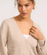 Cardigan corto de punto color beige claro, con escote en V, manga larga abullonada y cierre frontal de botones. Posee bolsillos delanteros tipo parche.