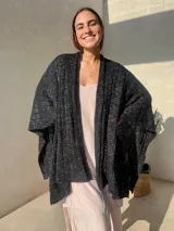 Ruana tejida en punto jersey, color negro con destellos brillantes de lurex. Presenta una etiqueta de cuero sintético con logo en la parte posterior.