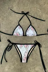 Bikini de dos piezas con corpiño triangular y bombacha regulable con lazos laterales. El estampado es floral en tonos rosados sobre fondo blanco, con ribete negro.