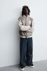 Cazadora estilo varsity o bomber de hombre, color gris oscuro con efecto ante, mangas en tono gris claro y puños, cuello y bajo con rib elástico a rayas blancas y grises. Presenta un parche con la letra 'R' en el pecho derecho y un bordado con texto en el pecho izquierdo.