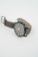 Reloj analógico con caja oscura y esfera marrón, con tres subesferas de cronógrafo. Correa de cuero marrón con costuras blancas visibles y hebilla metálica.