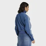 Campera Adidas modelo Firebird confeccionada en tejido denim de algodón. Presenta cierre frontal de cremallera, cuello alto, puños y ruedo en tejido rib elástico. Incluye las icónicas tres franjas en las mangas y el logo del trébol bordado en el pecho.