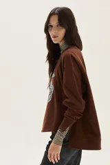 Buzo oversize de felpa color marrón con bordado frontal de una flor estilizada y la palabra "Muse" escrita en cursiva en color celeste.