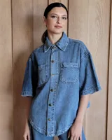 Sobrecamisa de denim soft en tono azul medio, con corte oversize, mangas cortas, cuello clásico y cierre frontal con botones metálicos. Presenta dos bolsillos frontales con solapa y un ruedo redondeado.