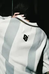 Chomba de manga larga con diseño de rayas verticales en color blanco y verde seco. Presenta cuello tipo polo con cierre de botones y parches decorativos en el pecho.