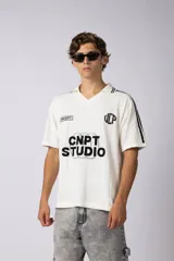 Remera tipo chomba de tejido de punto color blanco, con cuello en V y mangas cortas. Presenta rayas verticales sutiles en la trama y estampado gráfico frontal con la inscripción CNPT STUDIO y detalles en negro.