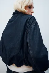 Cazadora de mezclilla azul oscuro, con cuello alto, manga larga, bolsillos ribeteados en el delantero y cierre frontal con cremallera y botones a presión ocultos bajo una solapa. Presenta costuras con pespuntes en contraste.