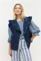 Chaleco de denim azul oscuro con volados estructurados en los hombros y el frente. Presenta cierre frontal de botones metálicos y bolsillos laterales.