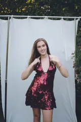 Vestido corto negro con escote en V profundo, estilo halter con tiras para atar al cuello. Presenta un diseño ajustado en el busto con tiras ajustables bajo el mismo que se cruzan y anudan en la espalda, y falda con volado en el ruedo con detalle de encaje.