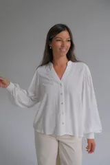 Blusa blanca de corte holgado con cuello en V, cierre frontal de botones y mangas largas abullonadas con puños elásticos. Presenta detalles bordados sutiles en la parte frontal y en las mangas.