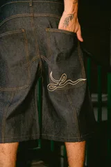 Bermuda de jean rígido color azul oscuro, corte baggy, con costuras contrastantes en color camel. Presenta un bordado estilizado en hilo blanco en el bolsillo trasero derecho.