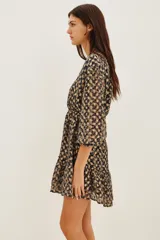 Vestido corto de escote en V cruzado, con mangas tres cuartos y cintura elástica. Presenta un diseño estampado con motivos geométricos en tonos oscuros y detalles metalizados, con un ruedo con volado.