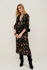 Vestido midi negro con estampado floral en tonos pastel y amarillos. Presenta escote pronunciado en V, mangas largas con puños anchos y detalles de bordado o pasamanería oscura en los bordes.